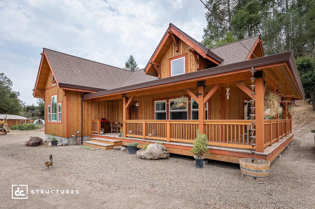 A rustic wooden house with a wraparound porch, surrounded by trees and a gravel path, embodies the charm of apartment barn kits. Featuring a steep roof and large windows, it offers a cozy, cabin-like appearance in its natural setting.