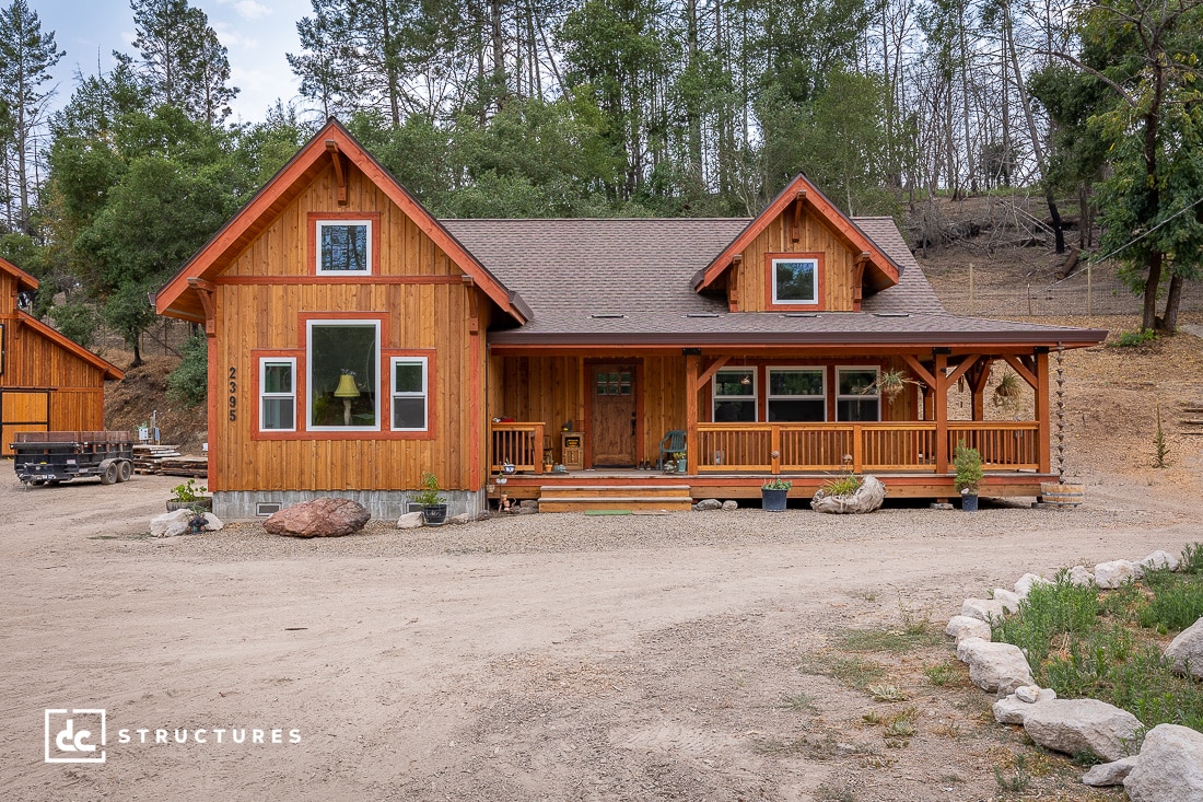 A rustic wooden cabin surrounded by trees, featuring a large covered porch with potted plants, evokes the charm of apartment barn kits. Set against a dirt driveway with forested hills as the backdrop, a small trailer is parked on the left, enhancing its quaint appeal.