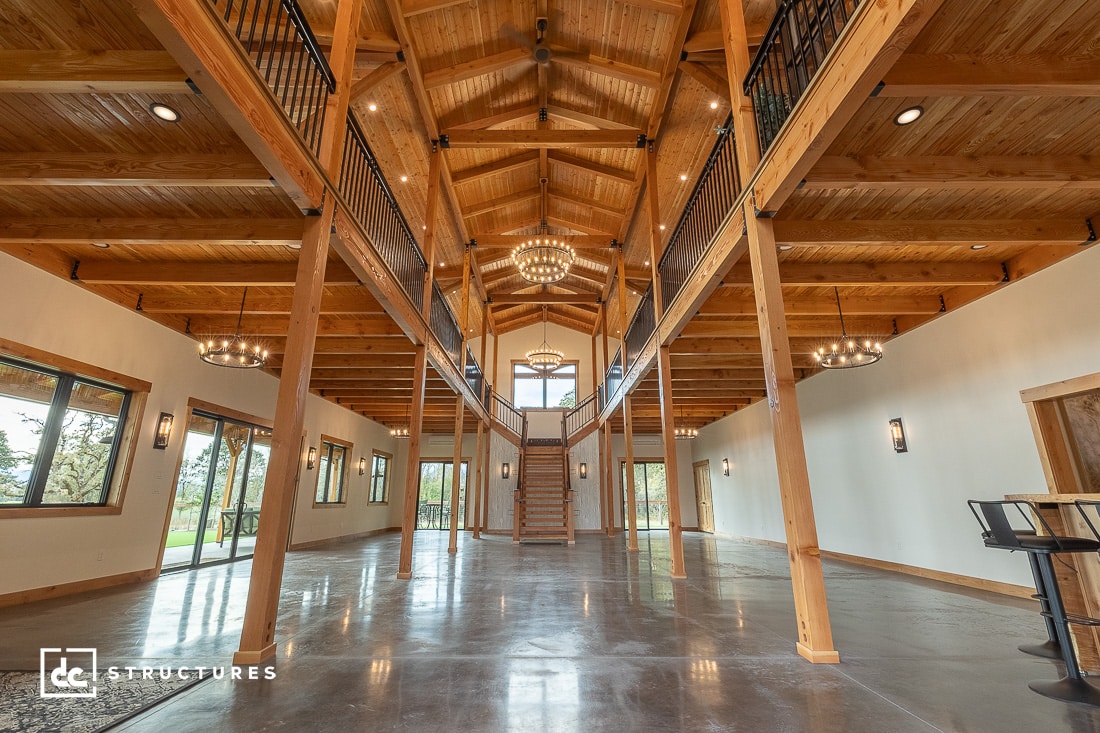 A spacious interior of a wooden barn with high ceilings, exposed beams, and chandeliers. Two staircases lead to an upper level. Large windows allow natural light in, and polished concrete floors reflect the warm tones of the wood.