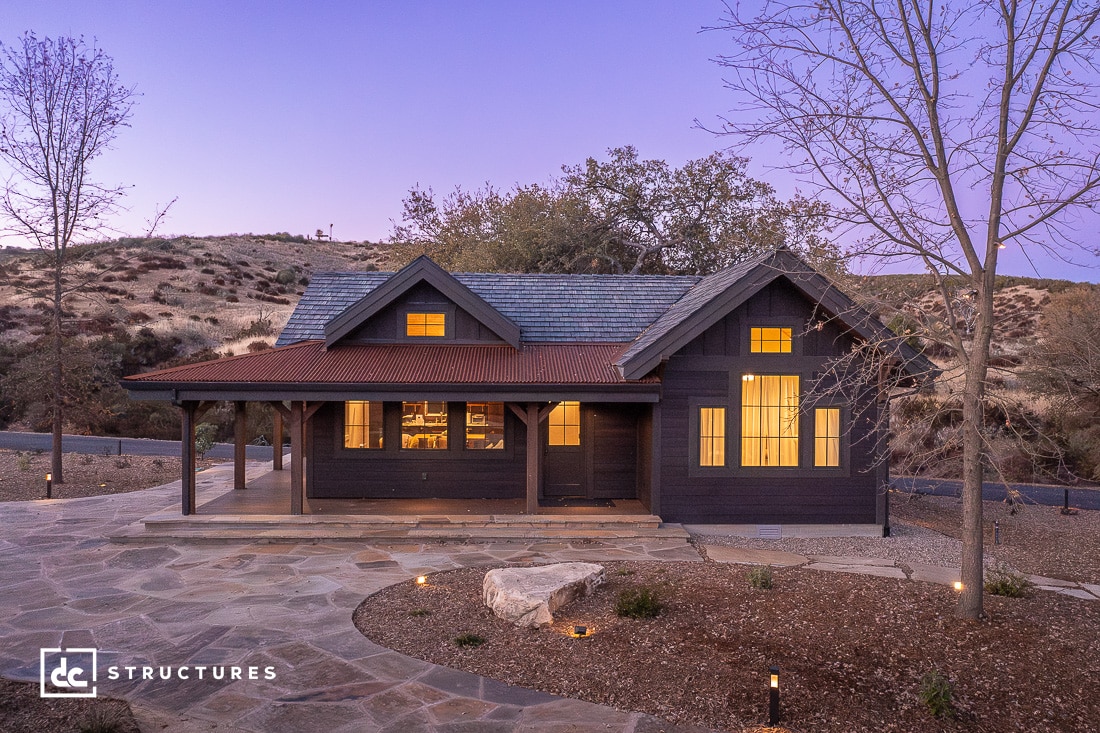 A rustic cabin with a wooden exterior and warm-lit windows stands at dusk, surrounded by a rocky landscape. Nearby, a barn adds charm to the scene, while a stone pathway leads to the entrance. A large tree enhances the setting as the sky takes on a soft purple hue.