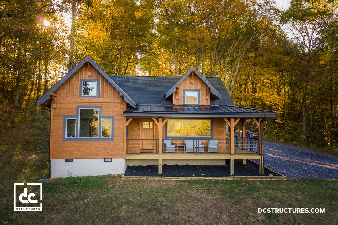 A rustic two-story wooden house with a porch and large windows, surrounded by autumn trees, evokes the charm of apartment barn kits. The dark roof complements the scene, while a gravel driveway leads to the entrance. The logo in the lower corners reads "DC Structures.