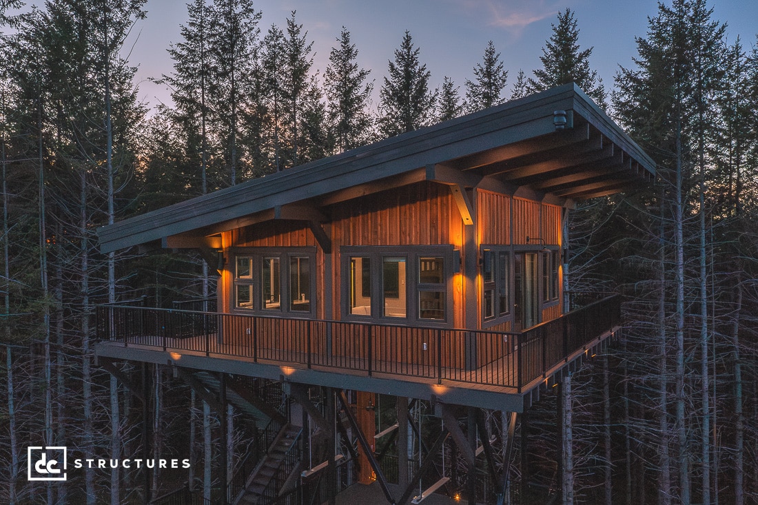 A modern treehouse with large windows and a slanted roof is elevated among tall trees. It features warm exterior lighting and a wraparound balcony. The logo "DC Structures" is in the bottom left corner.