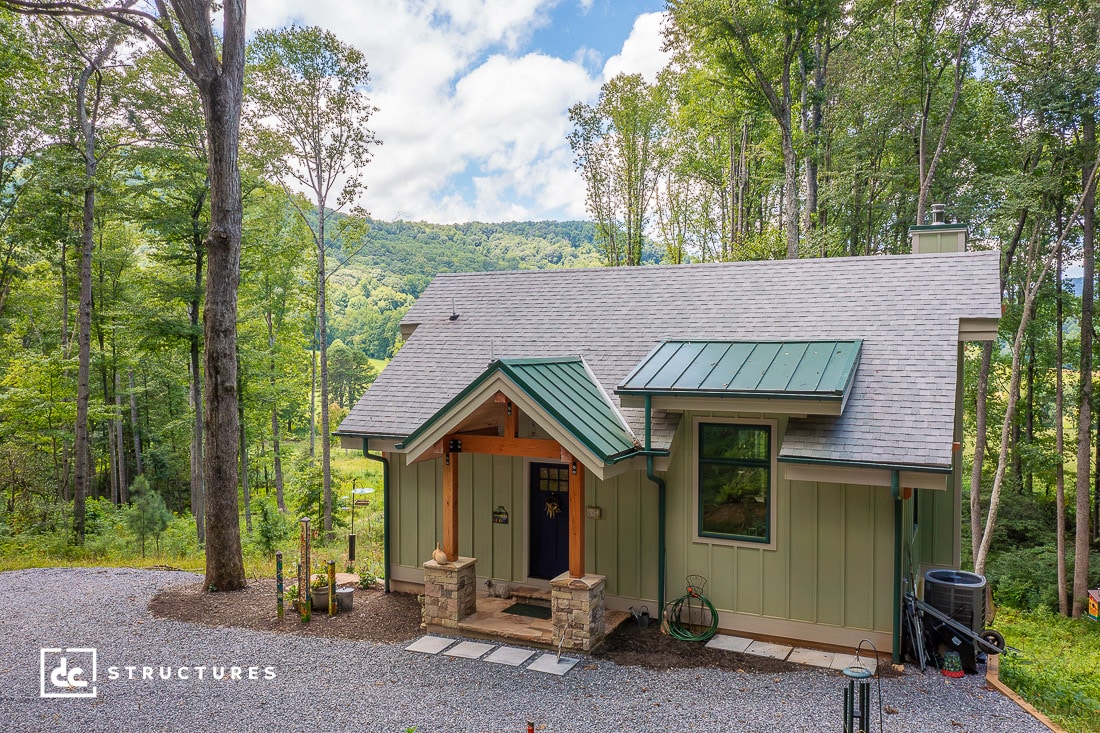 A small, modern cabin with a green metal roof and stone entrance is nestled in a lush forest setting. Tall trees surround the house, creating an inviting escape that blends rustic charm with elegance, much like apartment barn kits. A mountainous landscape peeks through under a partly cloudy sky.