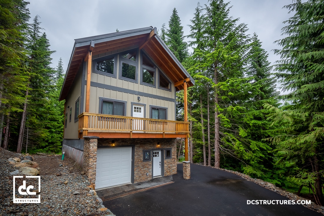 Nestled among tall pine trees, this two-story house boasts a stone foundation and wooden balcony, resembling a charming barn. The gray siding complements its rustic appeal. With several windows, a paved driveway, and an overcast sky above, it's as inviting as cozy barn kits.