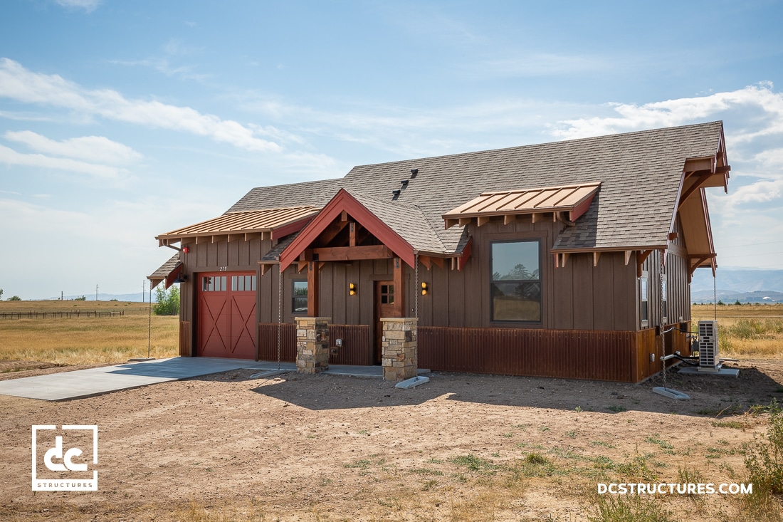 A small, rustic house with a gabled roof and wood siding features stone accents on the porch. With its red garage door and large windows, it resembles apartment barn kits. Nestled in a rural landscape, it boasts wide-open fields and distant mountains. Logo "dc structures" visible.