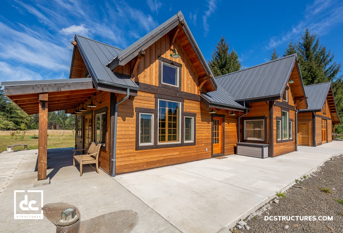 A modern wooden house with a gray metal roof under a clear blue sky resembles the simplicity of barn kits. The home features large windows and a spacious concrete patio with a bench. Tall trees and lush greenery envelop the property.