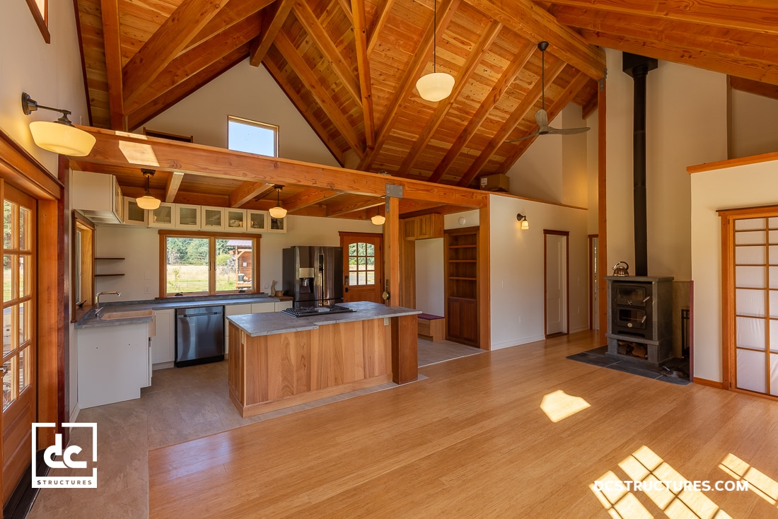 Spacious apartment with a high wooden ceiling and exposed beams, reminiscent of barn kits. It features a kitchen island and stainless steel appliances. A wood stove is positioned near large windows overlooking a lush green landscape, with polished wood floors enhancing its rustic charm.