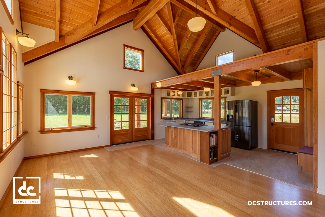 A spacious, sunlit room with wooden floors and high ceilings features a kitchen island with a stove, stainless steel appliances, and large windows. Reminiscent of modern barn aesthetics, the open space is inviting with exposed beams and warm lighting.