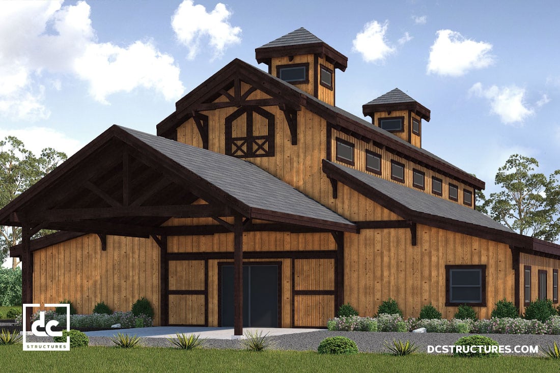 A large wooden barn-style building with a steep roof and multiple small windows. The structure has a prominent entrance under a covered porch. Surrounding the building is a neatly landscaped area with green plants and a clear blue sky above.