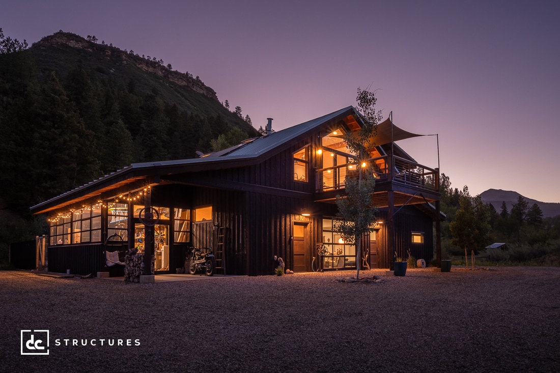 A modern barn-style house, crafted with elements from apartment barn kits, boasts large glass windows warmly illuminated at dusk. Nestled among forested hills and under a clear sky, it creates a serene evening ambiance.