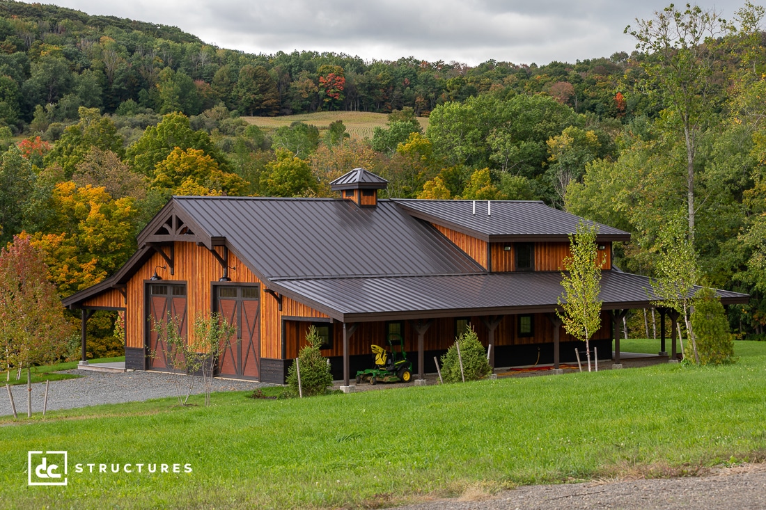 A modern wooden barn, reminiscent of stylish apartment barn kits, features a sleek black metal roof. Nestled in a lush landscape with trees and rolling hills under a cloudy sky, it boasts large sliding doors and a charming covered porch area.