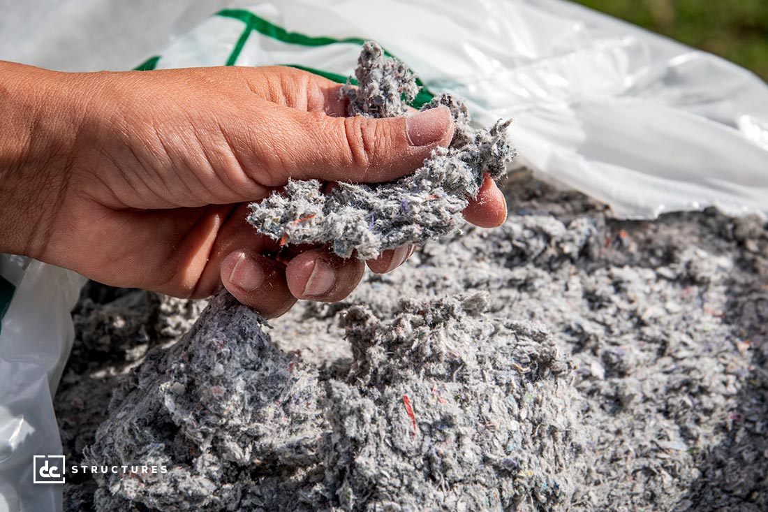 A hand holding a small amount of gray, fluffy insulation material, with more of the insulation in a large, open bag in the background. The texture appears soft and fibrous.