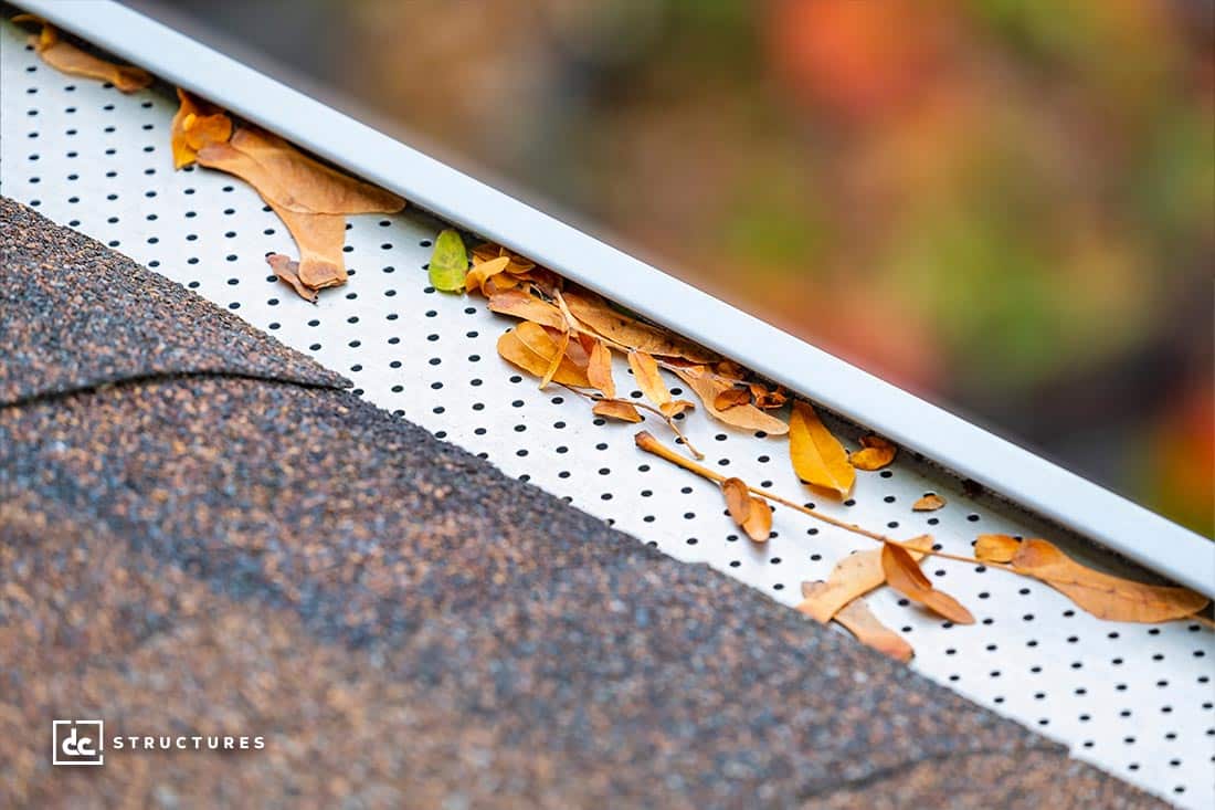Close-up of a gutter with a perforated guard partially covered by dry leaves. The roof has brown shingles, and the background is blurred with hints of orange and green, suggesting autumn foliage.