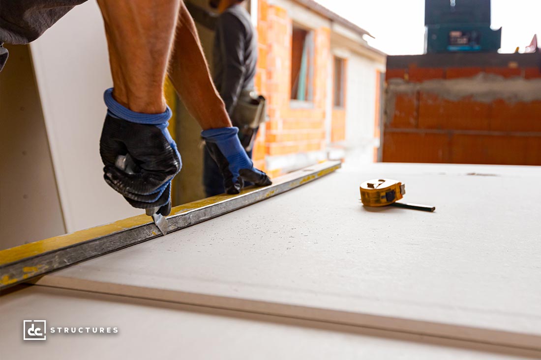 Two construction workers measuring and cutting drywall with a utility knife. They are using a yellow level tool for precision. A tape measure is on the surface. A partially constructed brick wall is visible in the background.
