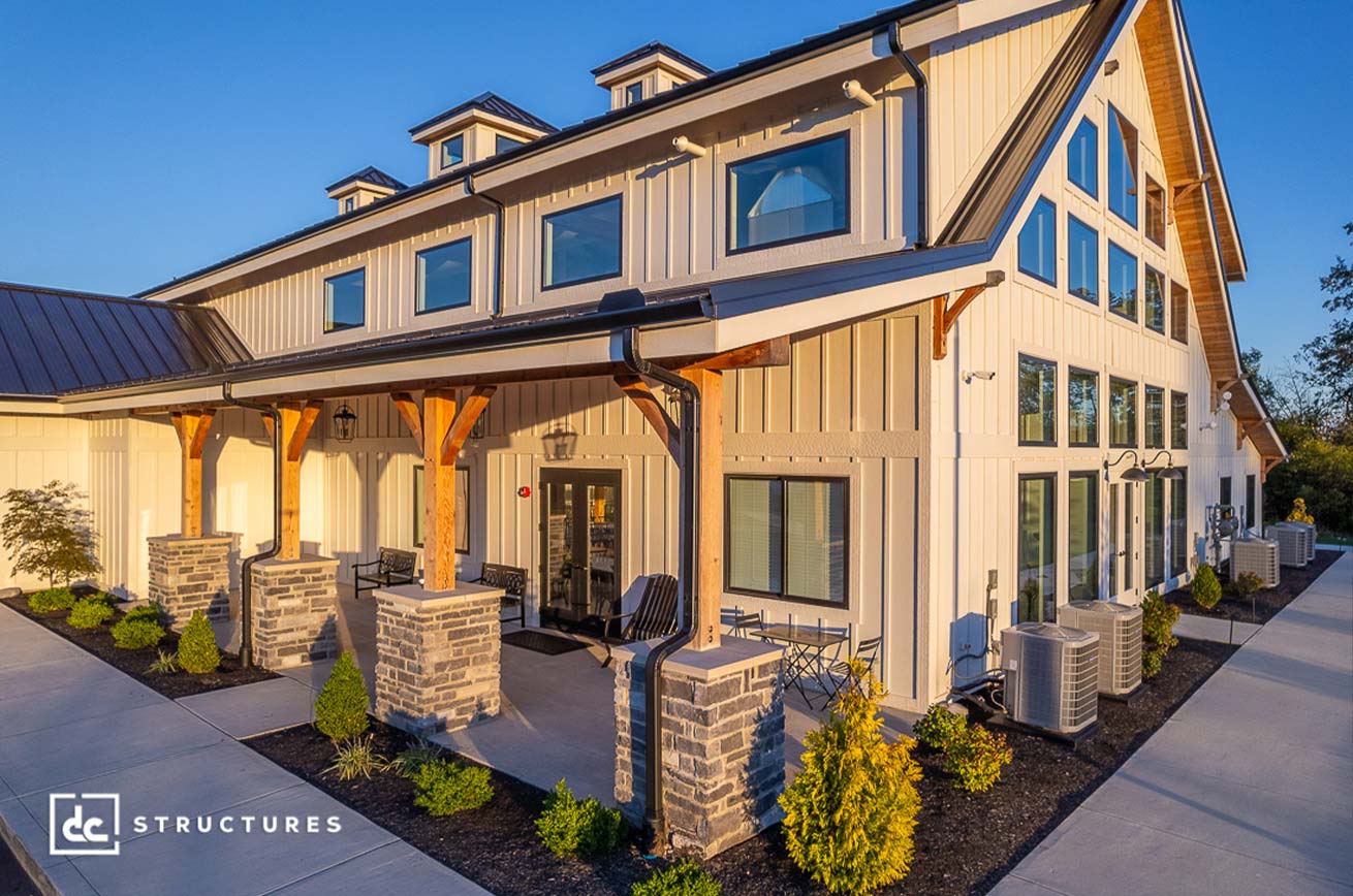 A modern building with a barn-style design features large windows, wooden beams, and a covered porch. It has a beige exterior, contrasting gray accents, and landscaped greenery along the pathway. The sky is clear and blue.