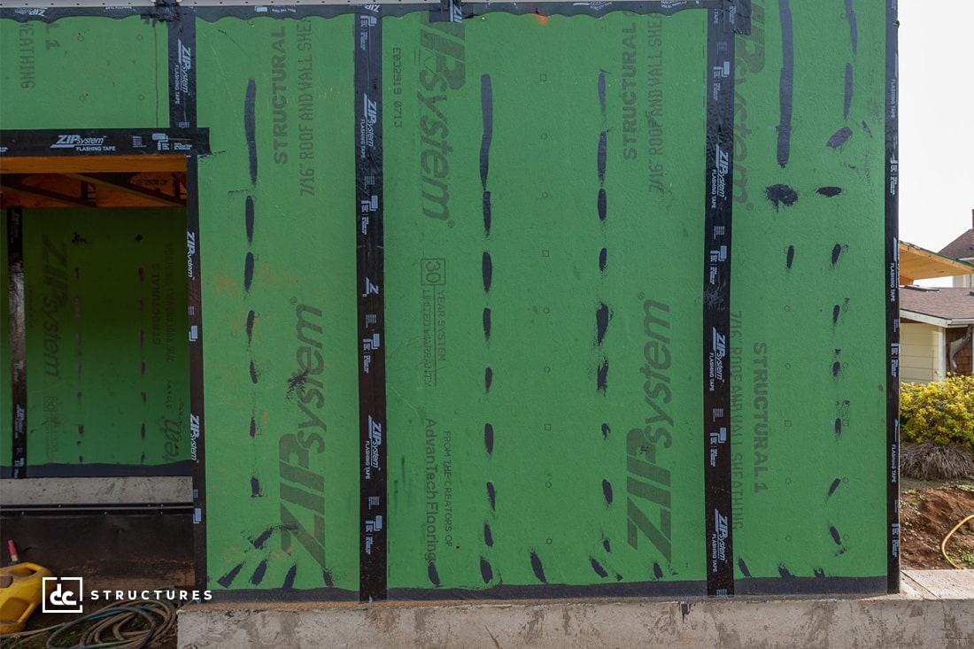 A partially constructed building with green sheathing labeled ZIP System and Structural 1. The exterior is visibly under construction, with black tape sealing the seams. A portion of the wooden frame is exposed on the left side.