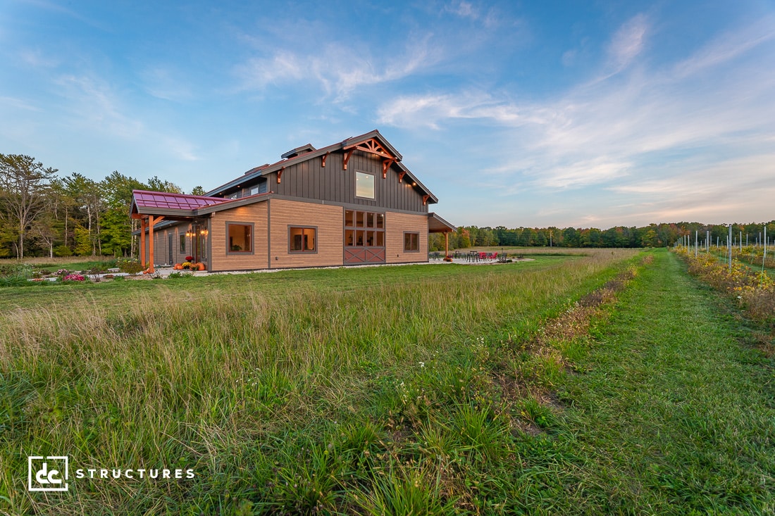 A modern barn-style house stands in a lush, open field with a clear blue sky above. The building features solar panels on the roof and a mix of wood and metal materials. Trees line the horizon, and a green pathway extends beside the structure.