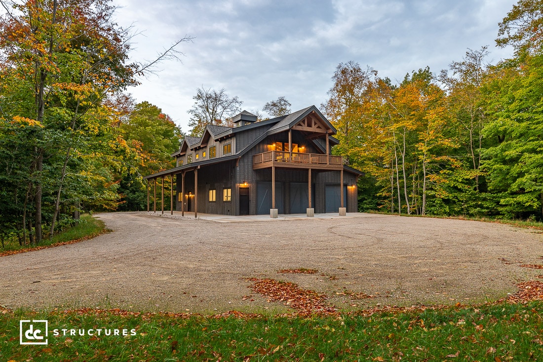 A modern two-story house with wooden accents surrounded by a forest. The house has a balcony and three garage doors. The trees display vibrant autumn colors. The sky is overcast, and fallen leaves decorate the gravel driveway.