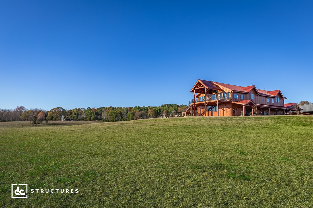 A large wooden house with a red roof sits on a vast green field under a clear blue sky. Trees are visible in the distance.