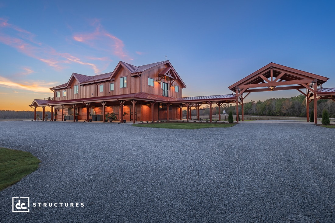 A large, rustic barn-style building with a gabled roof and wooden exterior sits under a clear blue and orange sunset sky. A covered walkway connects to a smaller structure on the right. Gravel driveway and open field surround the buildings.