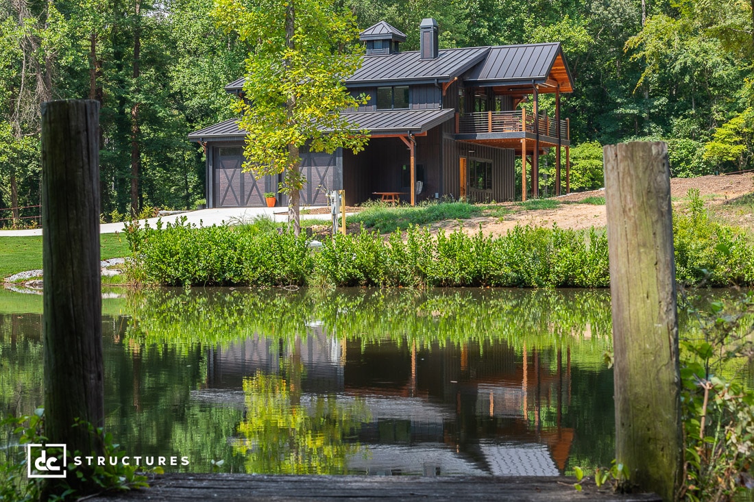 Modern two-story cabin with dark wood and metal accents, surrounded by lush greenery. The building is reflected in a calm pond in the foreground. A wooden fence and gravel path lead up to the house. Trees surround the serene setting.