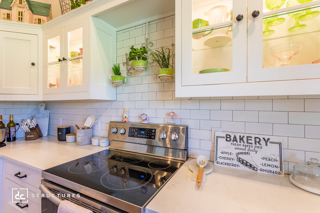 A bright kitchen featuring a stainless steel electric stove and white cabinets with glass doors displaying colorful dishes. The counter holds cooking utensils, potted plants, and a decorative bakery sign. Wine bottles are neatly arranged on the side.