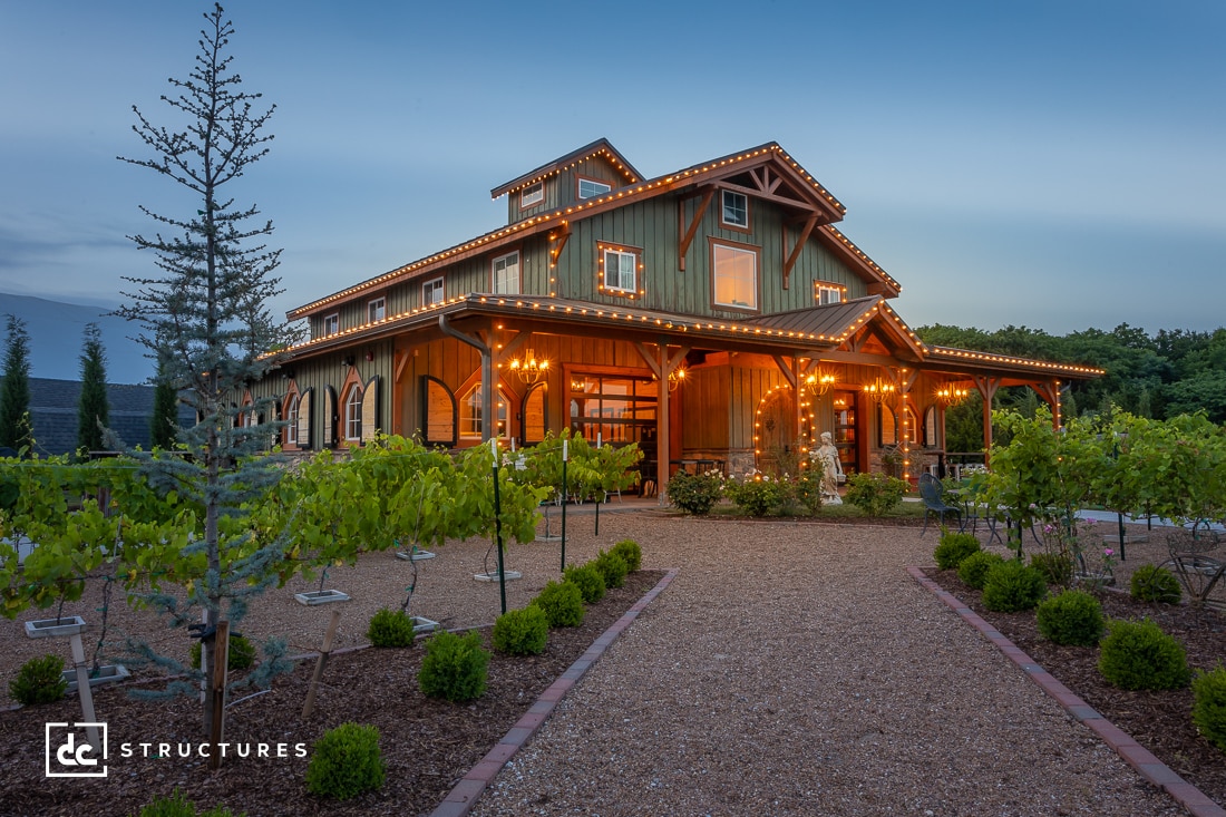 A rustic barn-style building with warm lights and green accents sits amid a landscaped garden with young trees and shrubs. The sky is a soft twilight blue, creating a serene and inviting atmosphere.