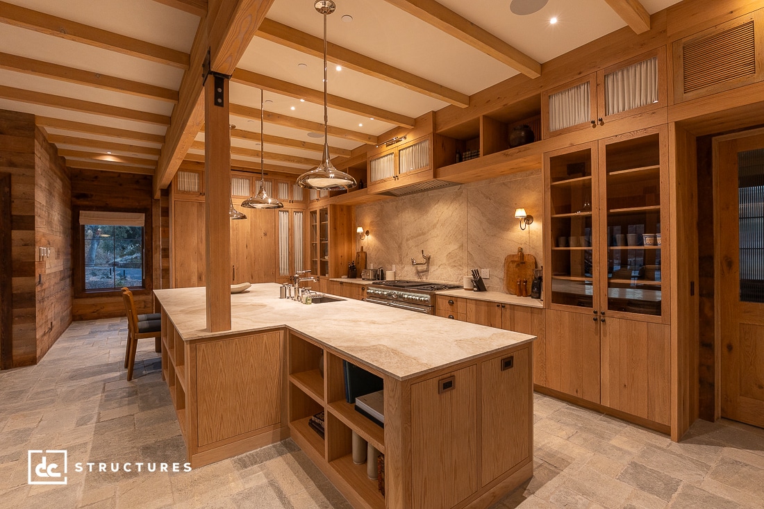 A spacious, rustic kitchen features wooden cabinets, a large central island with stone countertops, pendant lighting, and a stove against a stone backsplash. The floor is tiled, and warm lighting creates a cozy atmosphere.