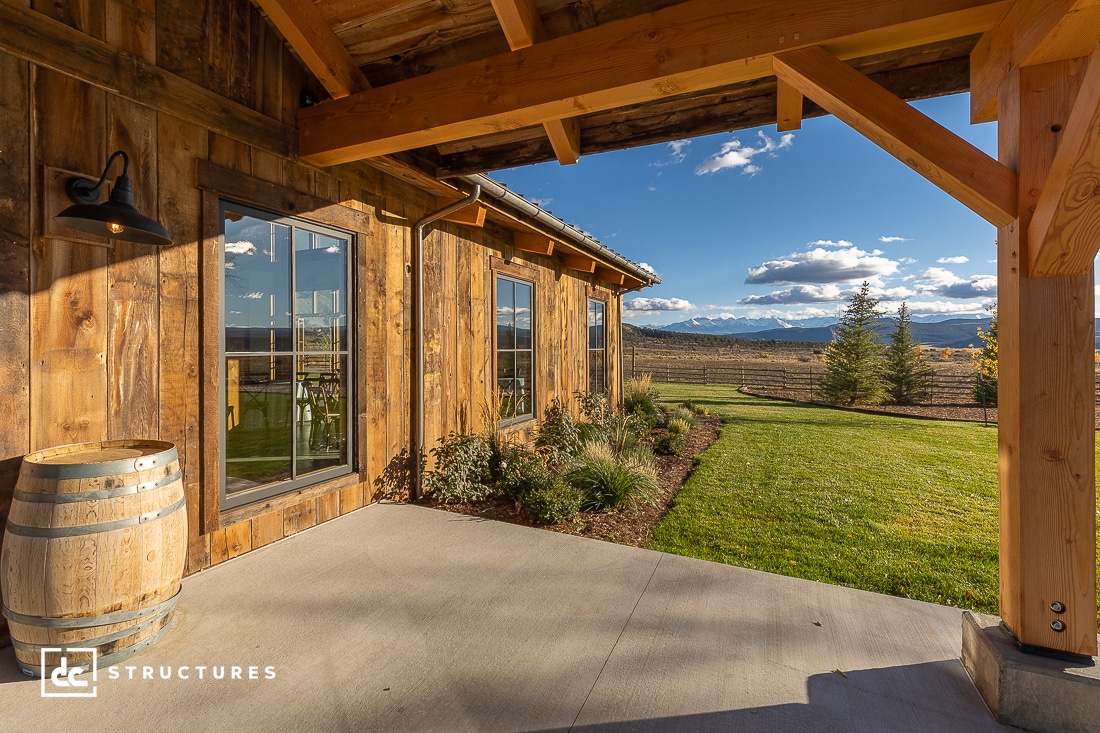 A rustic wooden building features large windows and a barrel on its porch. The porch overlooks a well-kept lawn with a view of distant mountains under a partly cloudy sky.