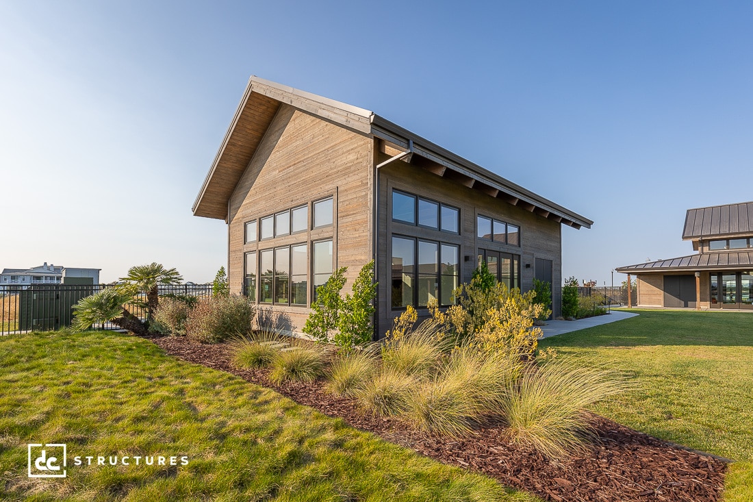 A modern, wooden house with large windows and a high-pitched roof sits on a landscaped lawn. Tall grasses and shrubs border the foreground, and there's a glimpse of another building in the background. The sky is clear and blue.