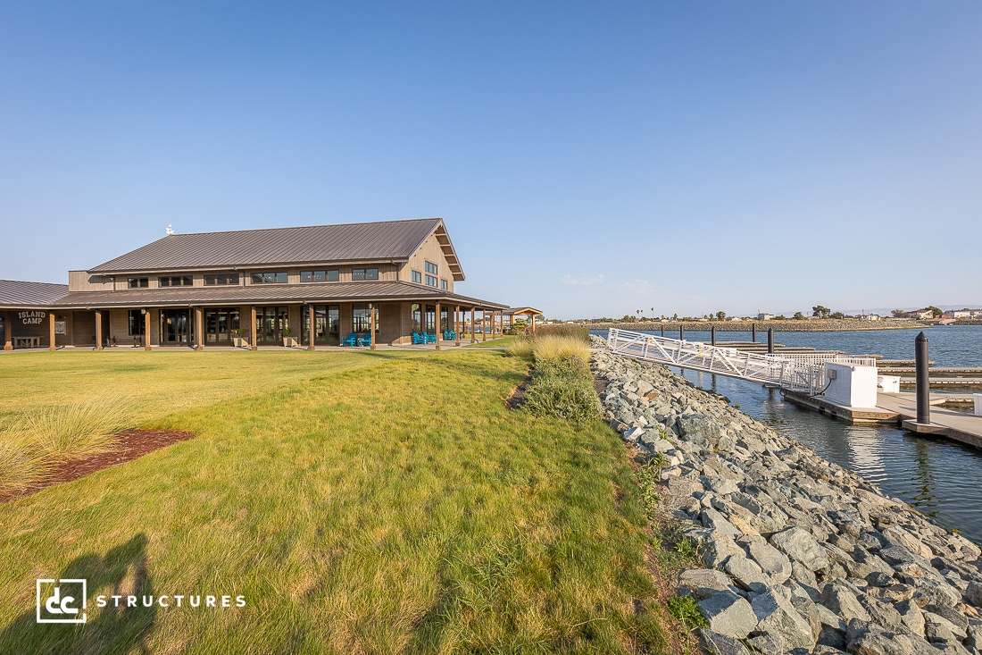 A modern building with large glass windows sits on a grassy area by the water. A rocky embankment leads to a dock extending over the calm water. The sky is clear, suggesting a sunny day.