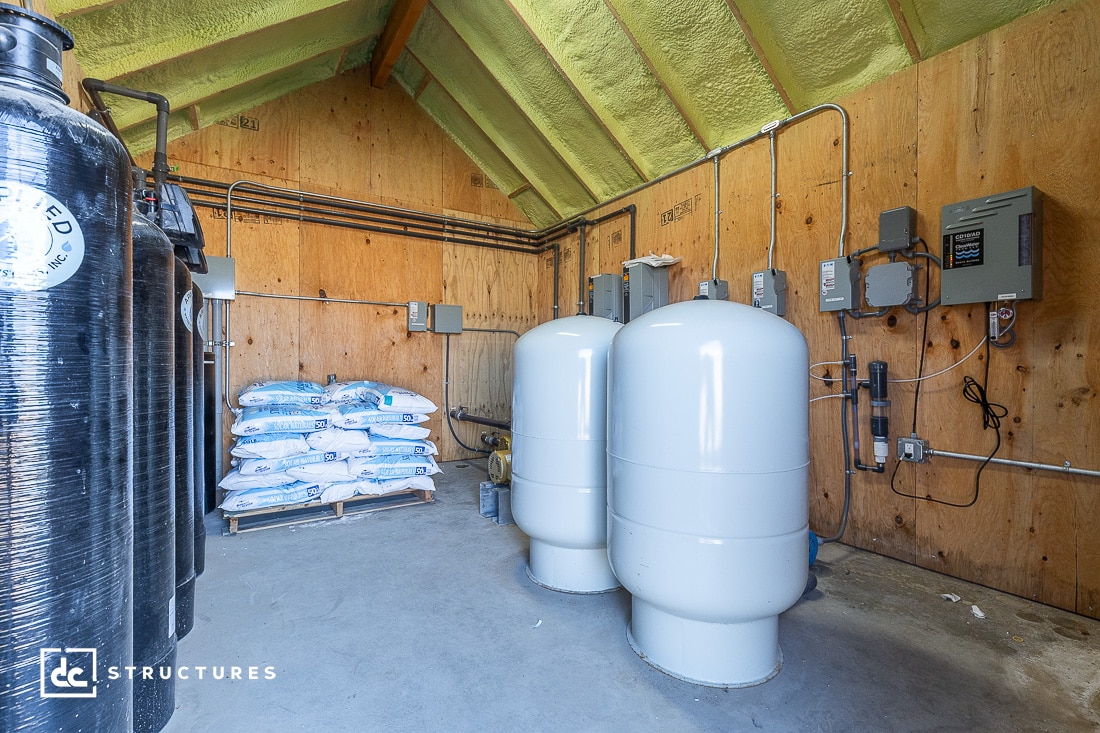 Interior of a wooden utility room with large water tanks, bags of water softener salt, and an array of electrical boxes on the walls. The ceiling is peaked and insulated with spray foam.