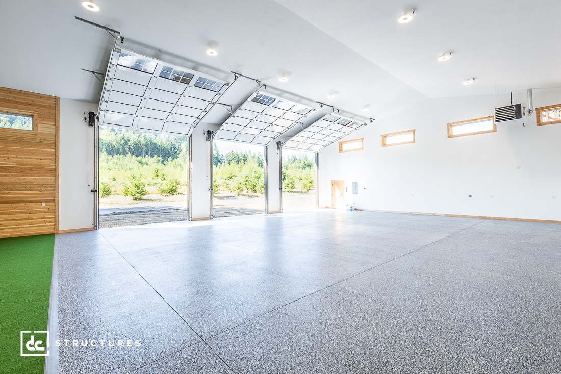 Spacious, modern garage or warehouse with gray flooring, white walls, large windows, and three open glass garage doors facing a view of trees outside. Natural light fills the clean, empty interior.