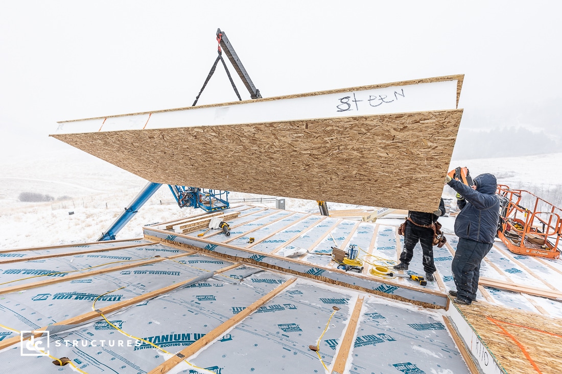 Construction workers install a large panel on a roof using a crane on a snowy day. The roof is covered with insulation material. Two men are guiding the panel into place. The landscape shows snow-covered hills in the background.