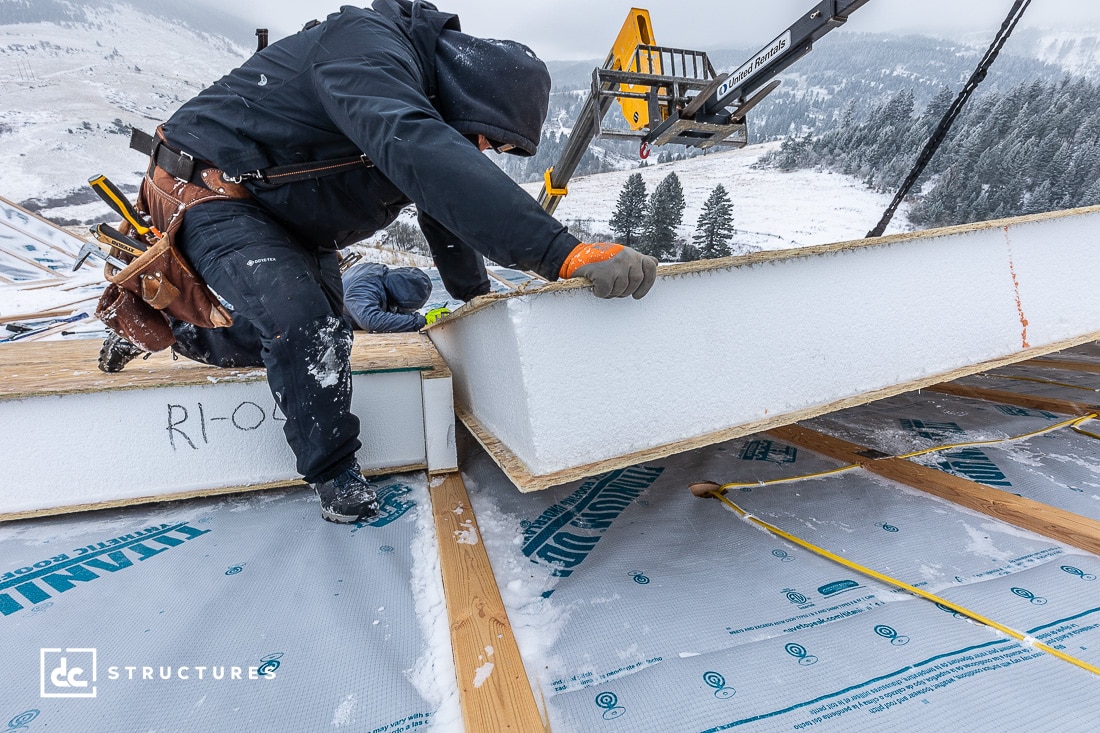 A construction worker in winter attire installs insulated panels on a snowy rooftop. The background features snow-covered trees and hills. A crane is lifting a panel in the distance. The scene conveys a cold-weather construction project.
