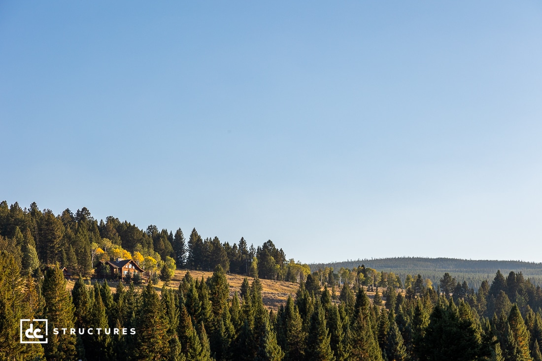 A scenic view of a forested hillside with a cabin nestled among trees. The sky is clear and blue, and the landscape is dotted with green and yellow foliage.