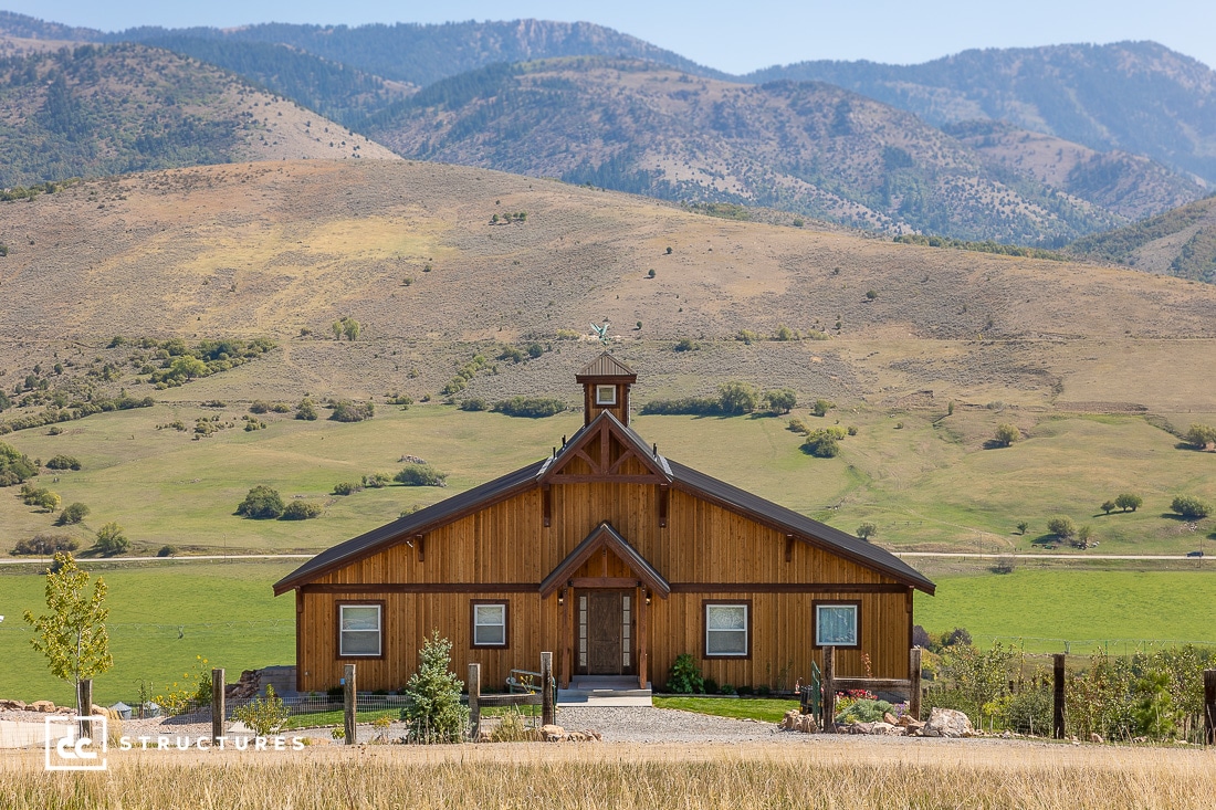 A wooden barn-style house stands in a green field with mountains in the background under a clear blue sky. The building has large front-facing windows and a small cupola on top. A gravel path leads to the front entrance.