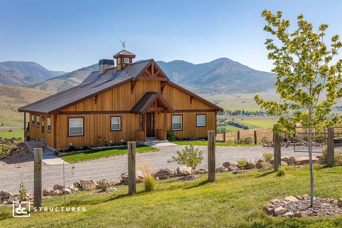 A wooden barn-style building with a triangular roof and cupola sits amidst a scenic rural landscape. Rolling hills and mountains are visible in the background. The foreground has a gravel path and a fenced area with a tree and rocks.