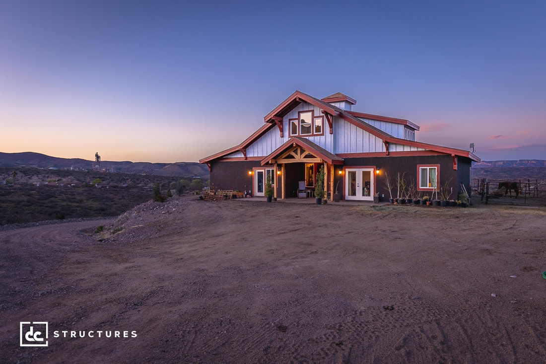 A rustic, two-story cabin with a metal roof stands in a vast, open landscape with a clear sky at dusk. Warm lights glow from the windows, and desert vegetation surrounds the building. A distant water tower is visible on the left.