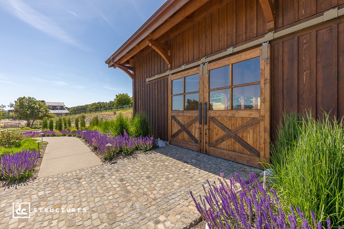 Wooden barn with large sliding doors, surrounded by a cobblestone path and blooming lavender plants. A concrete pathway curves towards the barn, with greenery and trees in the background under a clear blue sky.
