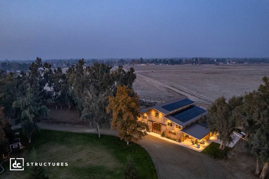 Aerial view of a large, modern barn-style structure surrounded by trees and expansive fields. The building is lit and has solar panels on the roof. A dirt driveway leads to the property. The sky is overcast.