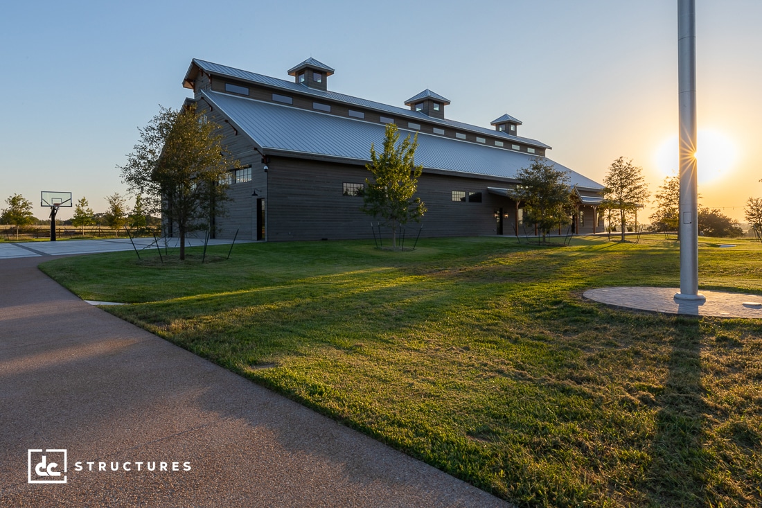 A modern barn-style building with a metal roof stands on a grassy area at sunset. The sky is clear, and the sun is setting on the horizon. A basketball hoop is seen on the left, and a flagpole on the right.
