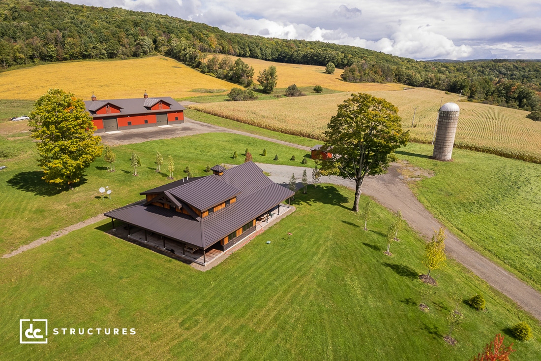 Aerial view of a countryside property with a modern barn-style house, red barn, silo, and expansive green lawns surrounded by fields and trees under a partly cloudy sky.