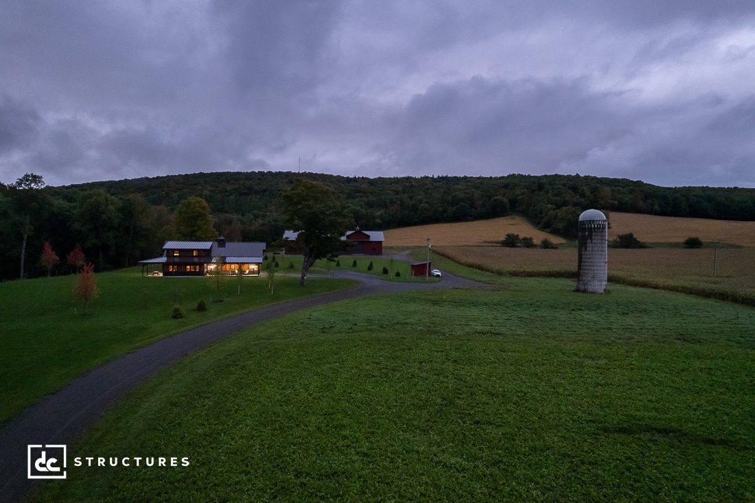 A scenic view of a rural landscape at dusk, featuring a farmhouse with lit windows, surrounded by fields and trees under a cloudy sky. A silo stands to the right near a winding driveway.