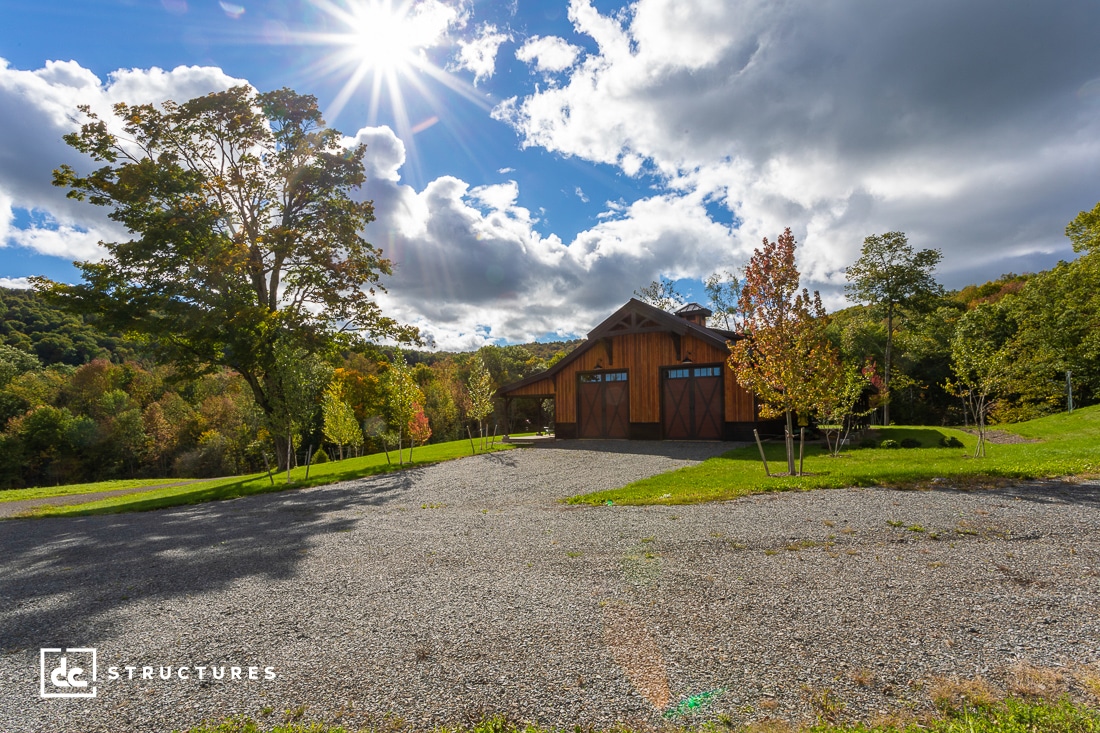 A rustic wooden barn with a large porch sits surrounded by green and autumn-colored trees under a bright, sunny sky. Gravel driveway leads to the barn. A small group of clouds floats overhead, and hills are visible in the background.
