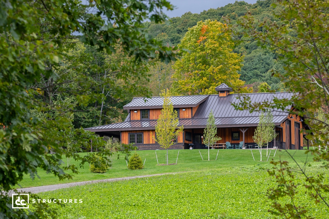 A wooden house with a metal roof is surrounded by lush green grass and trees. The structure blends with the natural landscape, and a small pathway leads to the house. The setting is peaceful, with a backdrop of green hills and a partly cloudy sky.