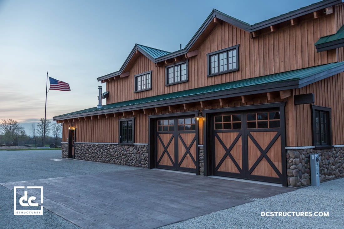 Two-story wooden building with a stone base and three large garage doors. An American flag is flying on a pole nearby. The sky is clear with a hint of sunset.