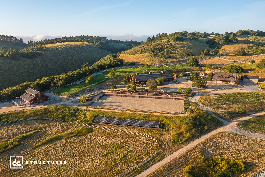 Aerial view of a rural landscape featuring a fenced equestrian arena and barn surrounded by green hills and trees. Various structures are scattered across the terrain, with winding dirt roads connecting them. The sky is clear with soft clouds in the distance.