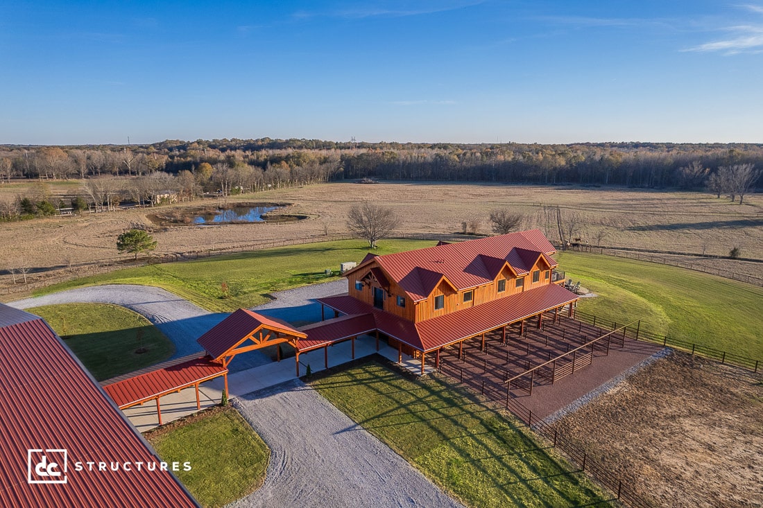 Aerial view of a large country-style house with red roofs, surrounded by green lawns and fields. The house has a driveway and a covered walkway. Nearby is a pond with surrounding trees under a clear blue sky.