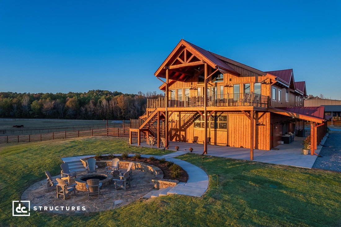 Large wooden barn-style home with a spacious upper deck and outdoor seating area surrounding a fire pit. The house is set against a backdrop of fields and trees, under a clear blue sky.